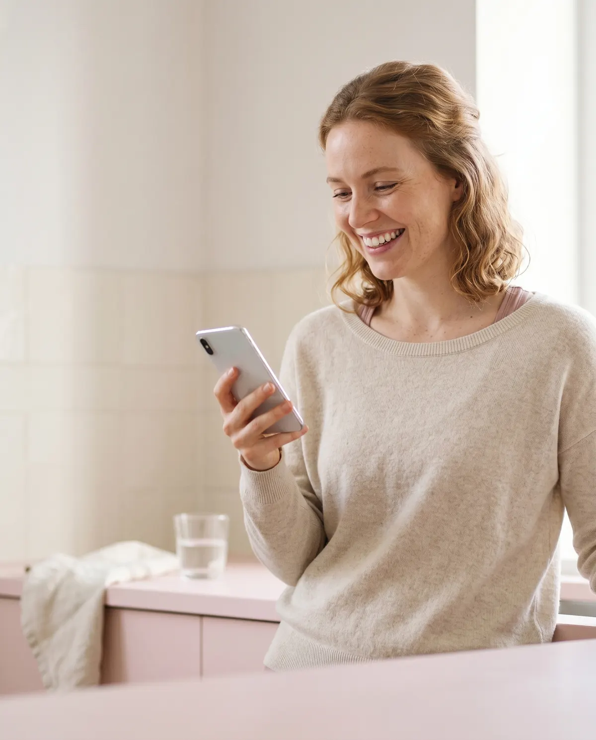 Woman in a cream sweater smiling at her phone in a soft pink kitchen during a standard telehealth consult